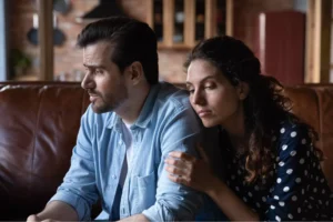 A couple sits close together on a couch, looking distressed and withdrawn, symbolizing the emotional process of healing from infidelity.