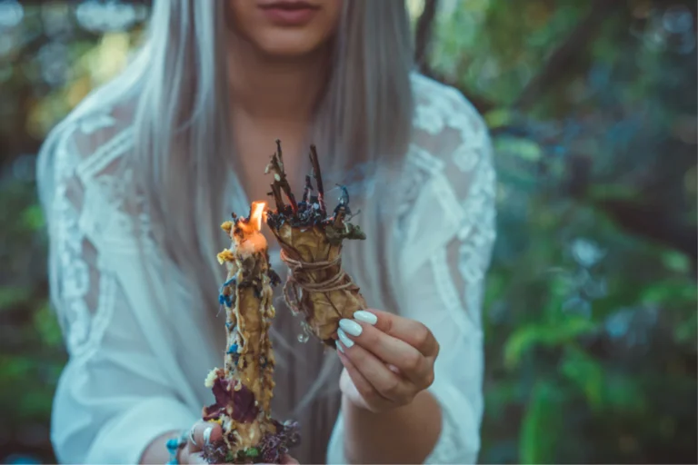 Young woman with long gray hair holds a burning sage bundle outdoors, referencing modern Wiccan practices.